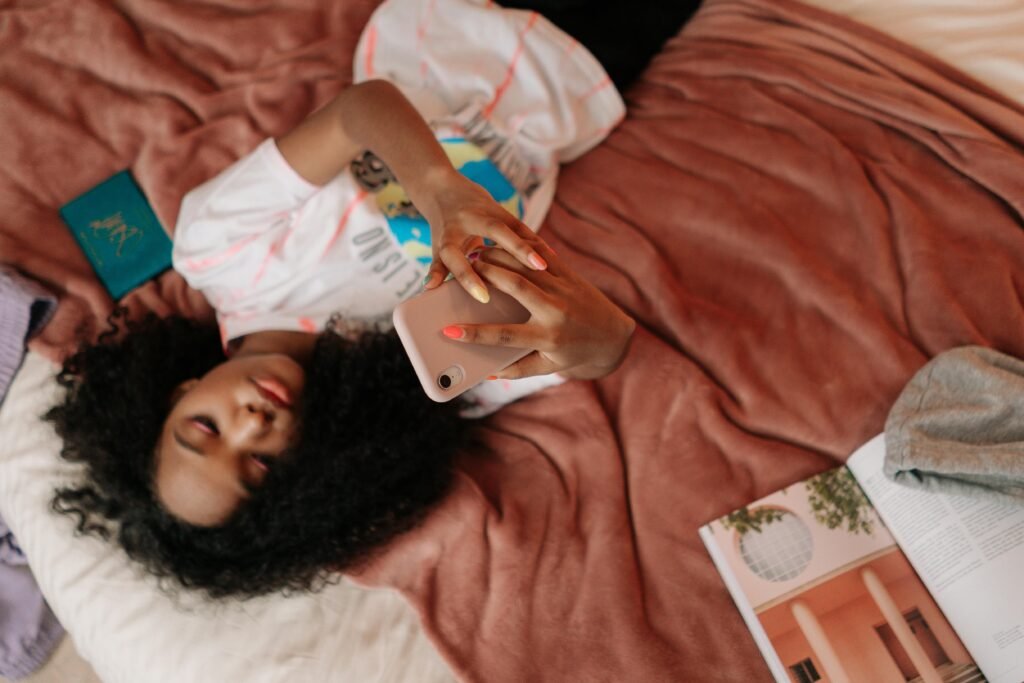 Teen girl on bed using smartphone, surrounded by book and blanket. Cozy indoors setting.